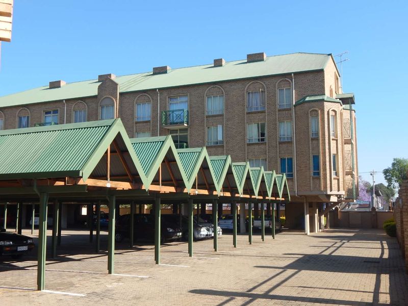 Exterior view of a multi-story brick apartment building featuring a covered parking area with a distinctive green corrugated metal roof structure.