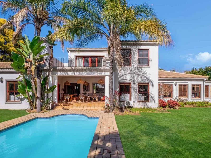 Rear view of a two-story residence featuring a swimming pool, paved surround, covered patio, and an upper-level balcony set within a landscaped garden.