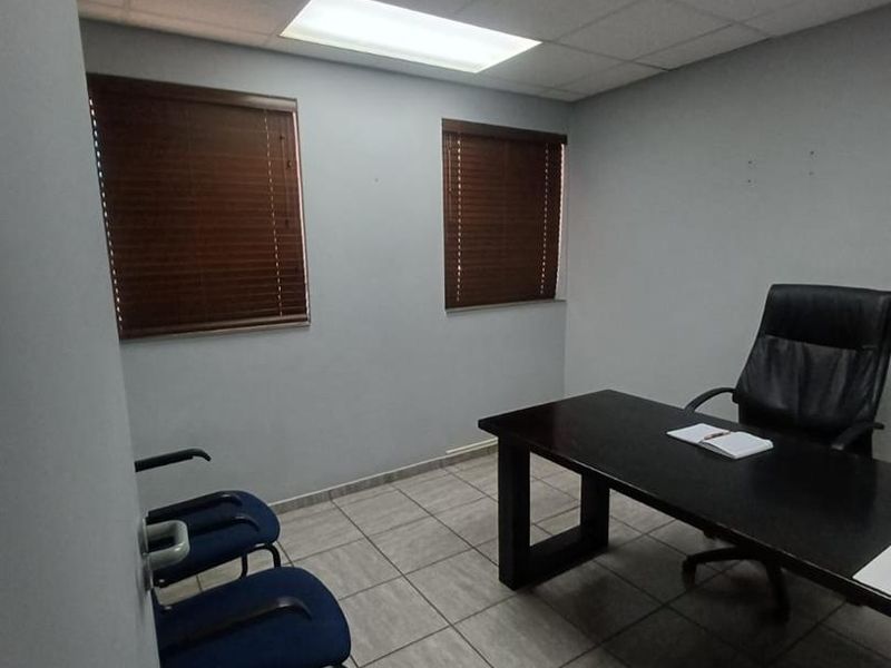 A functional office space featuring tiled flooring, a suspended ceiling with recessed lighting, and two windows fitted with venetian blinds.
