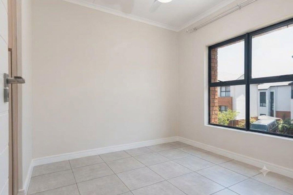 A bright, empty bedroom featuring neutral tiled flooring, a ceiling fan, and large aluminium-framed windows providing natural light.
