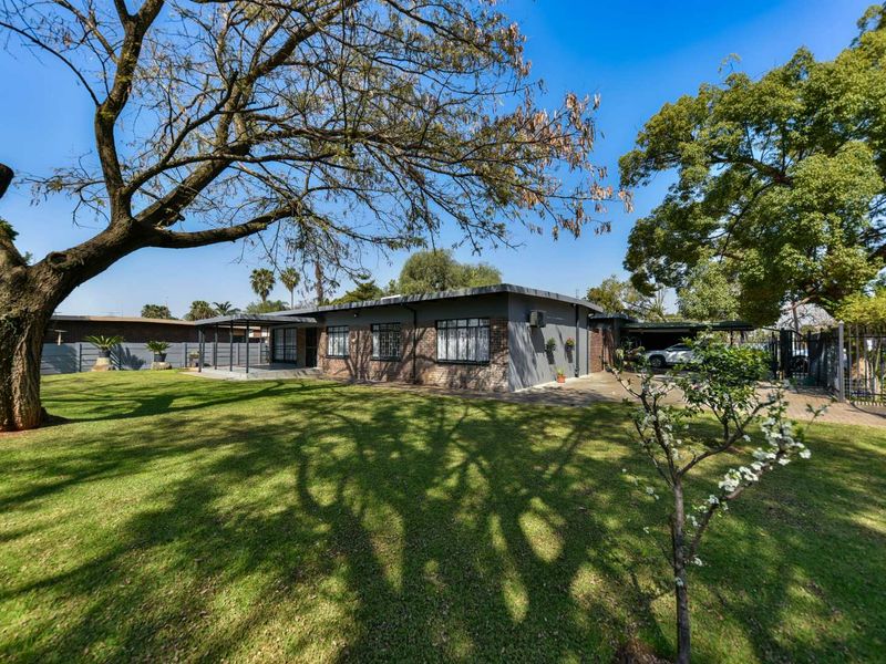 A spacious rear garden featuring a well-maintained lawn, mature trees, a covered patio area, and a paved driveway with secure perimeter fencing.