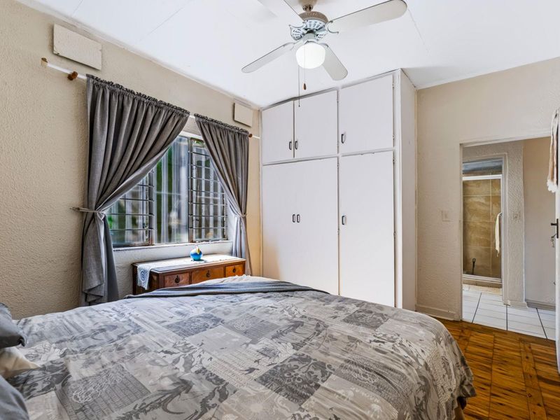 Bedroom featuring built-in cupboards, a ceiling fan, and wooden parquet flooring, with an adjoining bathroom visible through the doorway.