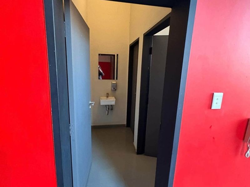A functional industrial-style bathroom facility featuring a wall-mounted basin, mirror, and soap dispenser, accessed through a bold red-painted entryway.