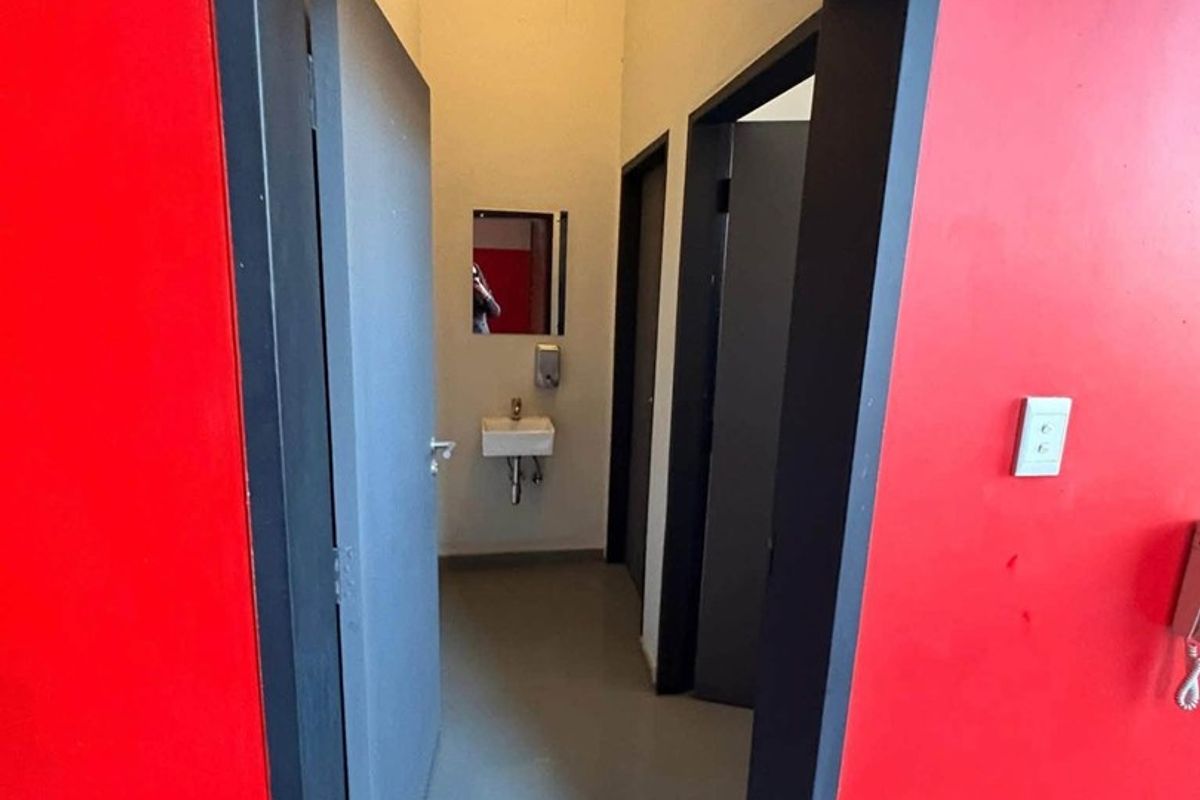 A functional industrial-style bathroom facility featuring a wall-mounted basin, mirror, and soap dispenser, accessed through a bold red-painted entryway.
