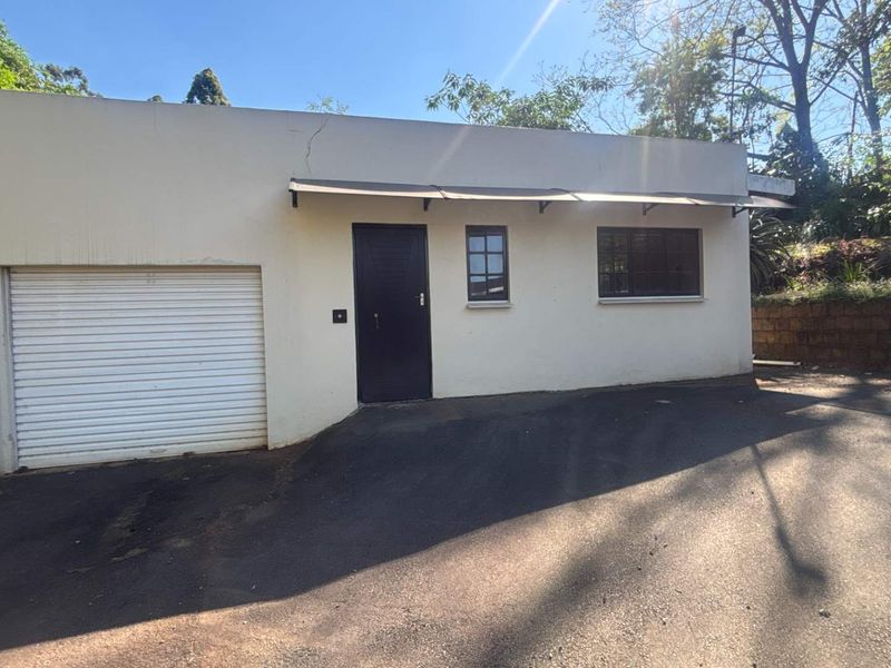 Front exterior view of a cottage featuring a roller shutter garage door, metal awning, and asphalt driveway with visible wall plaster cracking.