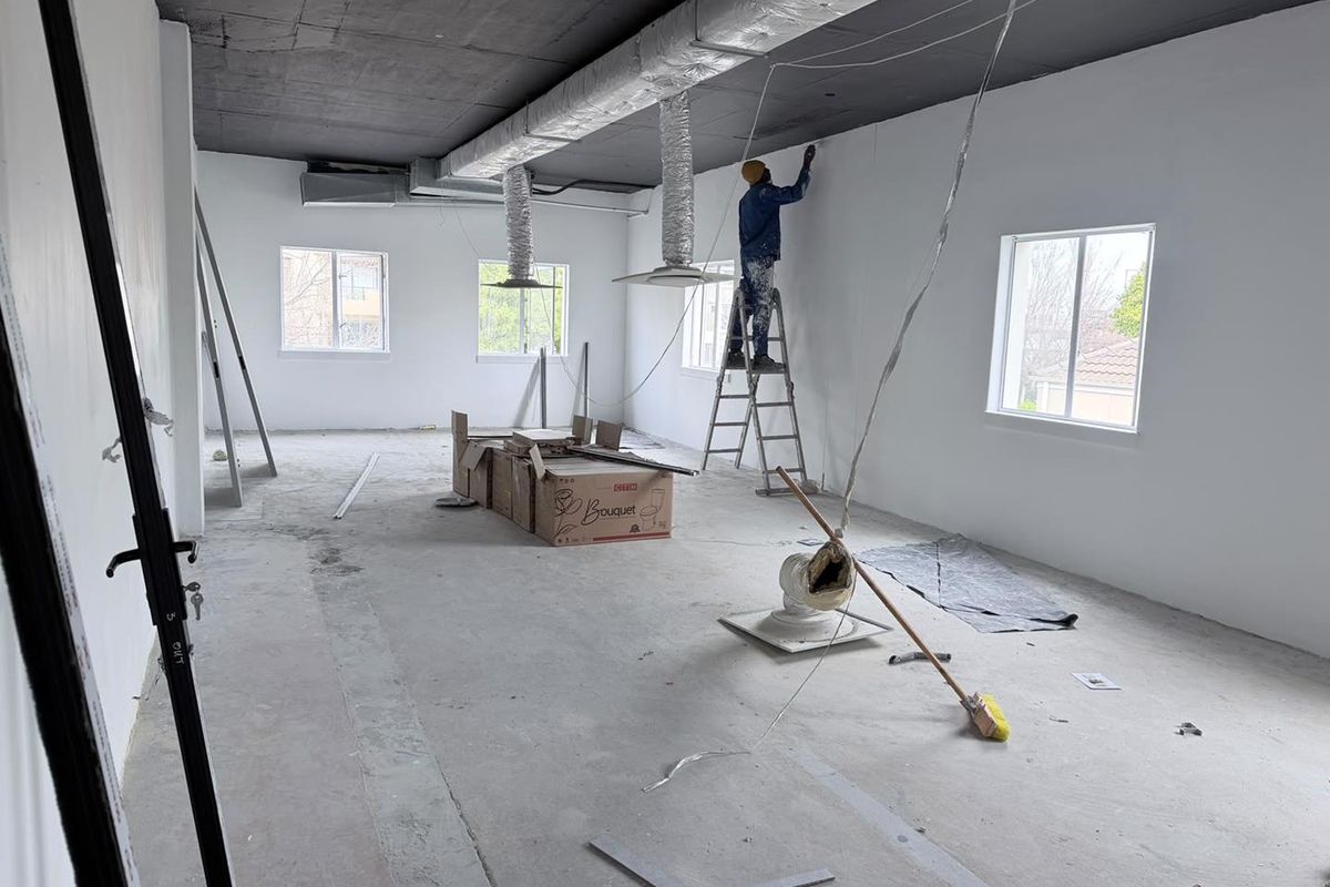 An unfinished office space featuring exposed ceiling ductwork, concrete flooring, and large windows, currently undergoing renovation and painting.
