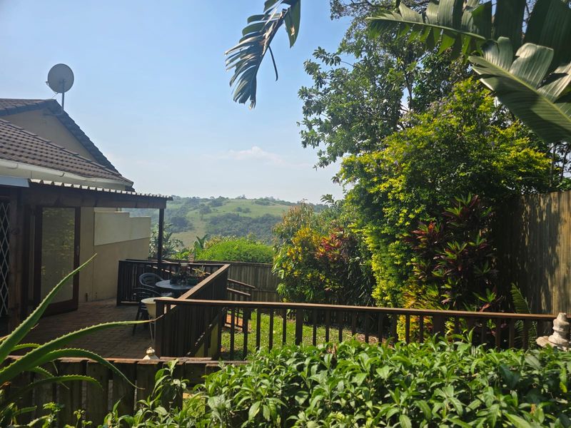 A raised timber deck and paved patio area featuring a pergola, overlooking a lush garden and scenic rolling hills.