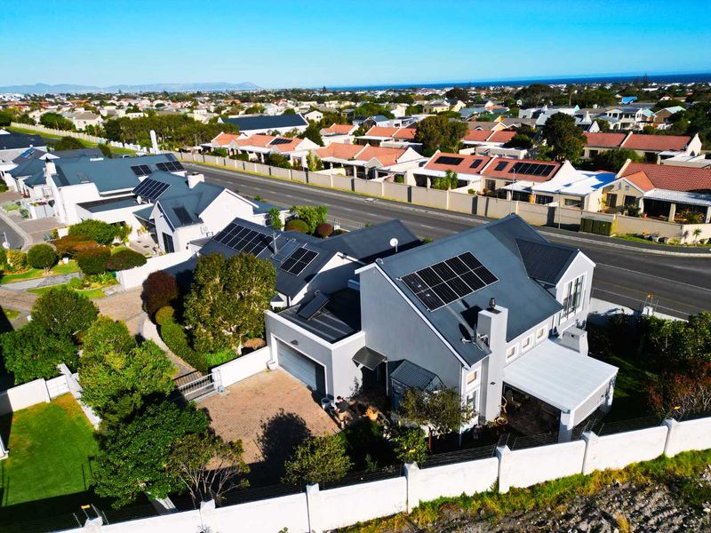 Aerial view of a residential property featuring a metal roof with solar panels, a paved driveway, a garage, and a secure perimeter wall.