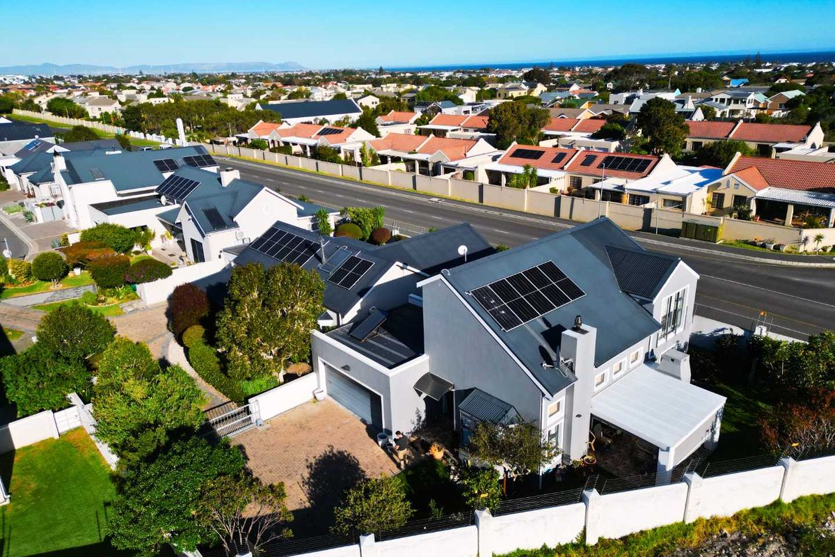 Aerial view of a residential property featuring a metal roof with solar panels, a paved driveway, a garage, and a secure perimeter wall.