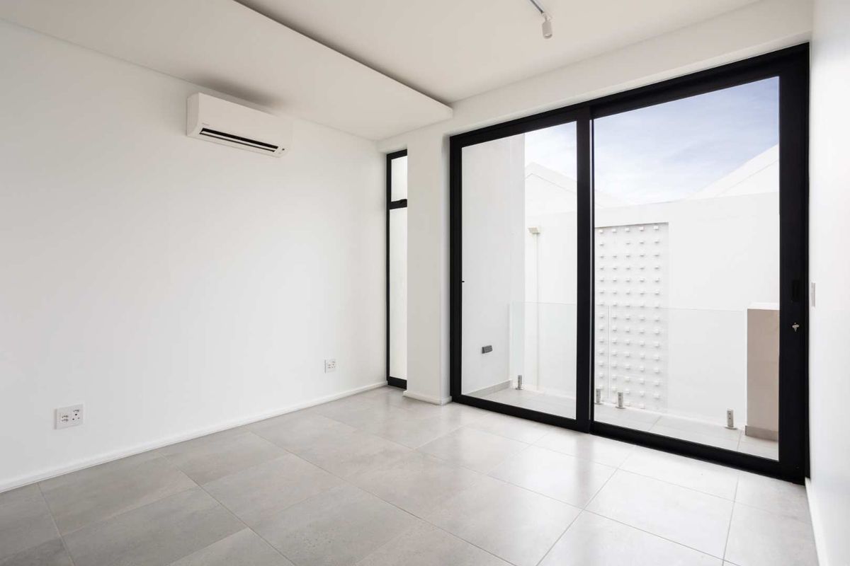A bright, modern bedroom featuring neutral tiled flooring, a split-system air conditioner, and large sliding glass doors opening to a private balcony.