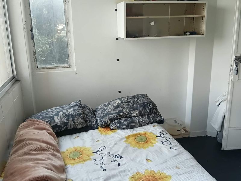 A compact bedroom featuring a wall-mounted storage unit and a window, requiring cosmetic updates to the wall finishes and window frame.