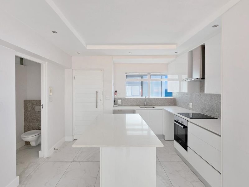 Modern, minimalist kitchen featuring a central island, sleek white cabinetry, stone countertops, integrated appliances, and high-quality tiled flooring.