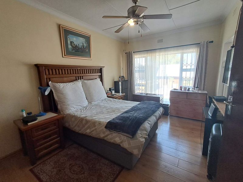 A well-lit bedroom featuring laminate flooring, a ceiling fan, and large windows with security gates, offering a comfortable and functional living space.