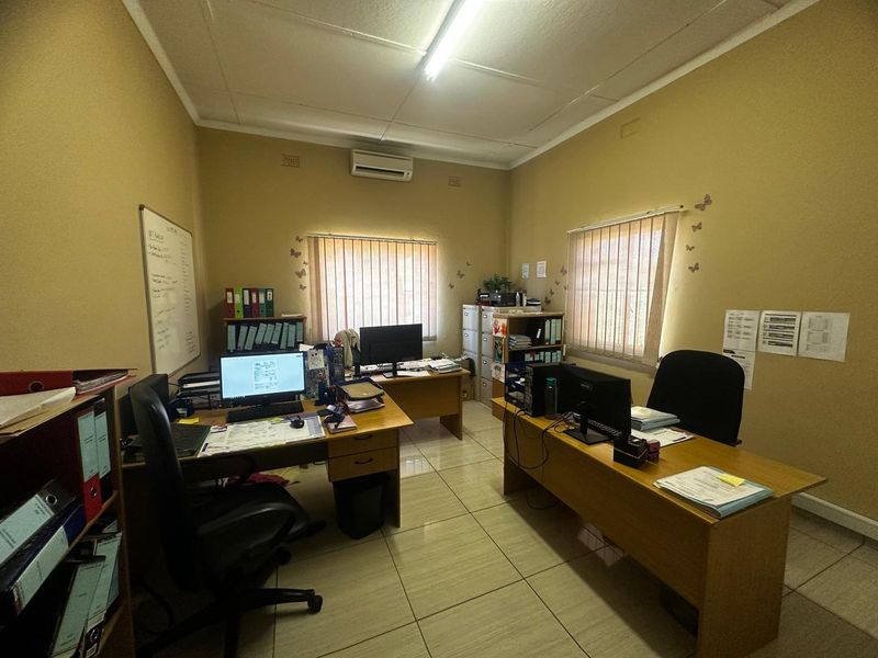 A functional office space featuring tiled flooring, a wall-mounted air conditioning unit, and dual windows with vertical blinds.