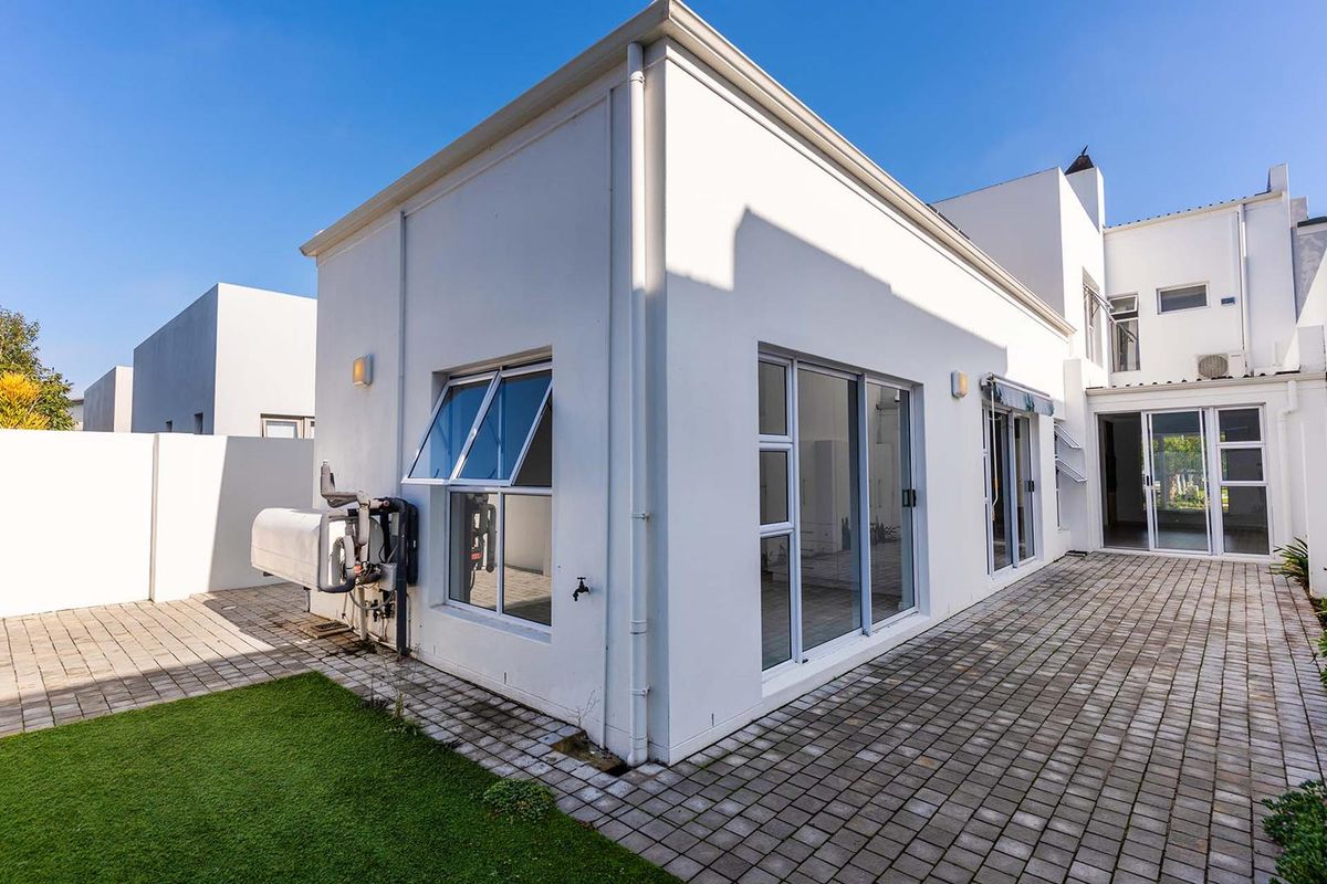 Rear exterior view of the property featuring paved patio area, white rendered walls, aluminium window frames, and a wall-mounted geyser unit.