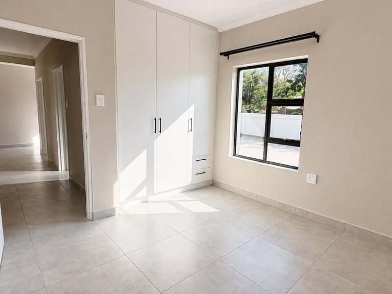 A bright, empty bedroom featuring neutral tiled flooring, built-in cupboards, and a large window allowing for ample natural light.
