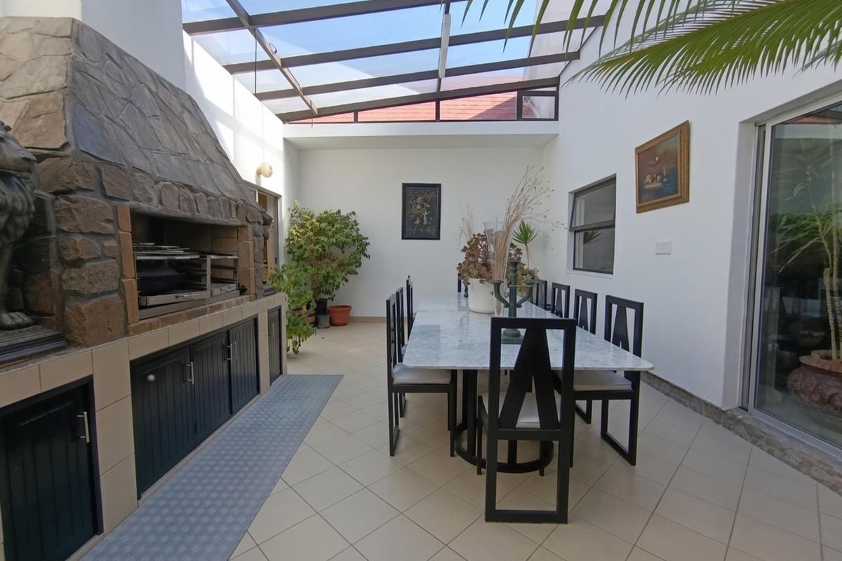 A bright, covered patio featuring a built-in stone braai, tiled flooring, and a glass roof, perfect for outdoor dining and entertaining.