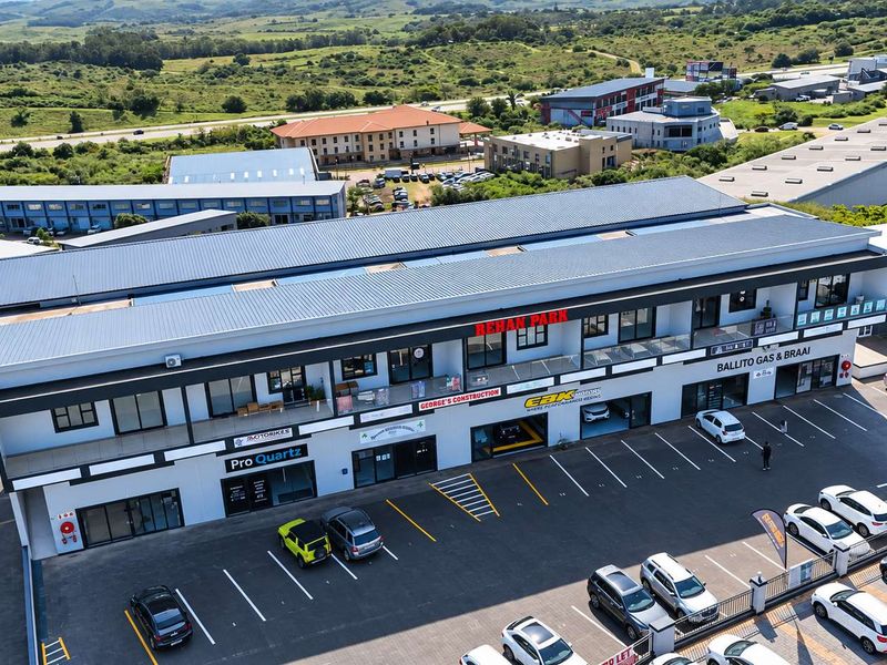 Aerial view of a modern multi-level commercial retail park featuring a metal roof, ground-level storefronts, upper-level balconies, and a paved parking area.
