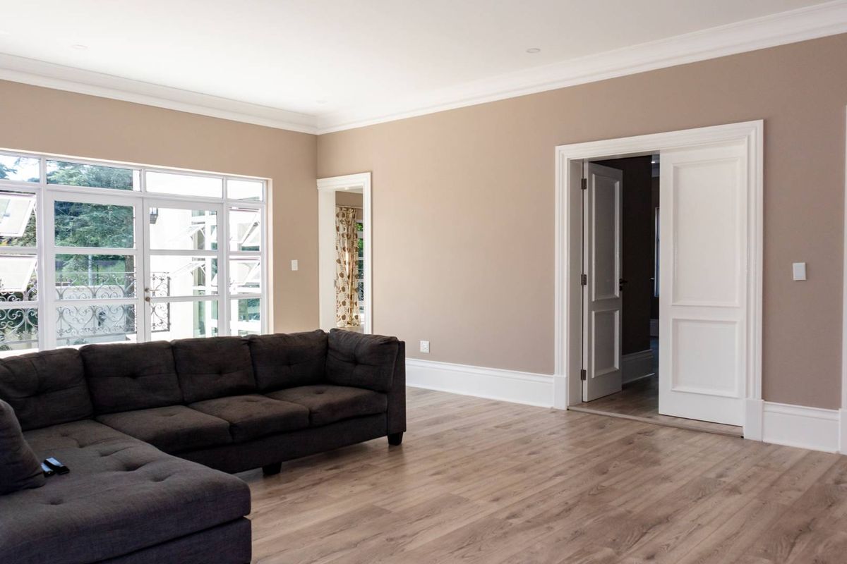 Spacious living room featuring light-toned laminate flooring, neutral wall finishes, decorative cornice, and large multi-pane windows providing natural light.