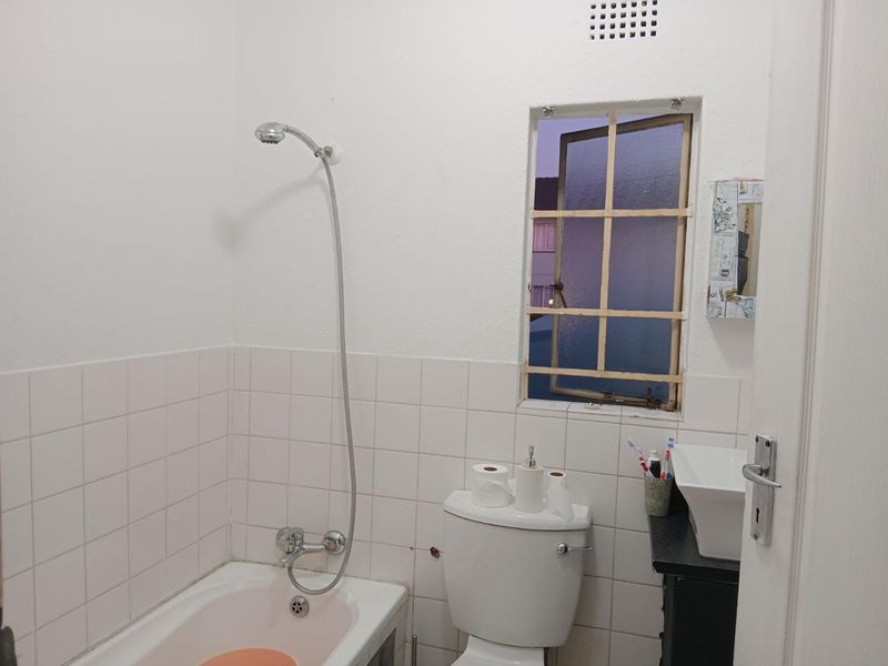 A compact bathroom featuring a bathtub with shower attachment, a toilet, and a vessel sink, requiring some cosmetic updates to the window frame.