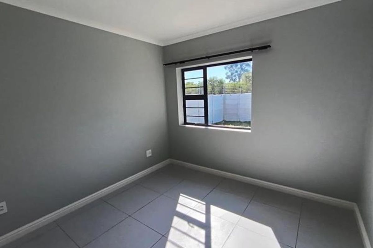 A clean, empty bedroom featuring neutral grey walls, large format floor tiles, and an aluminium-framed window with a curtain rod.
