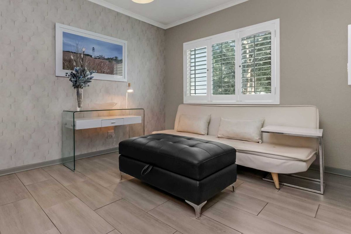 A well-maintained bedroom featuring neutral textured wallpaper, modern tiled flooring, and white plantation shutters providing natural light control.