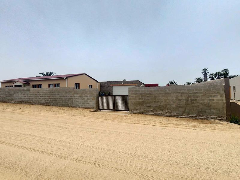 Front view of the property featuring a perimeter wall, metal gate, and garage access from a sandy road.