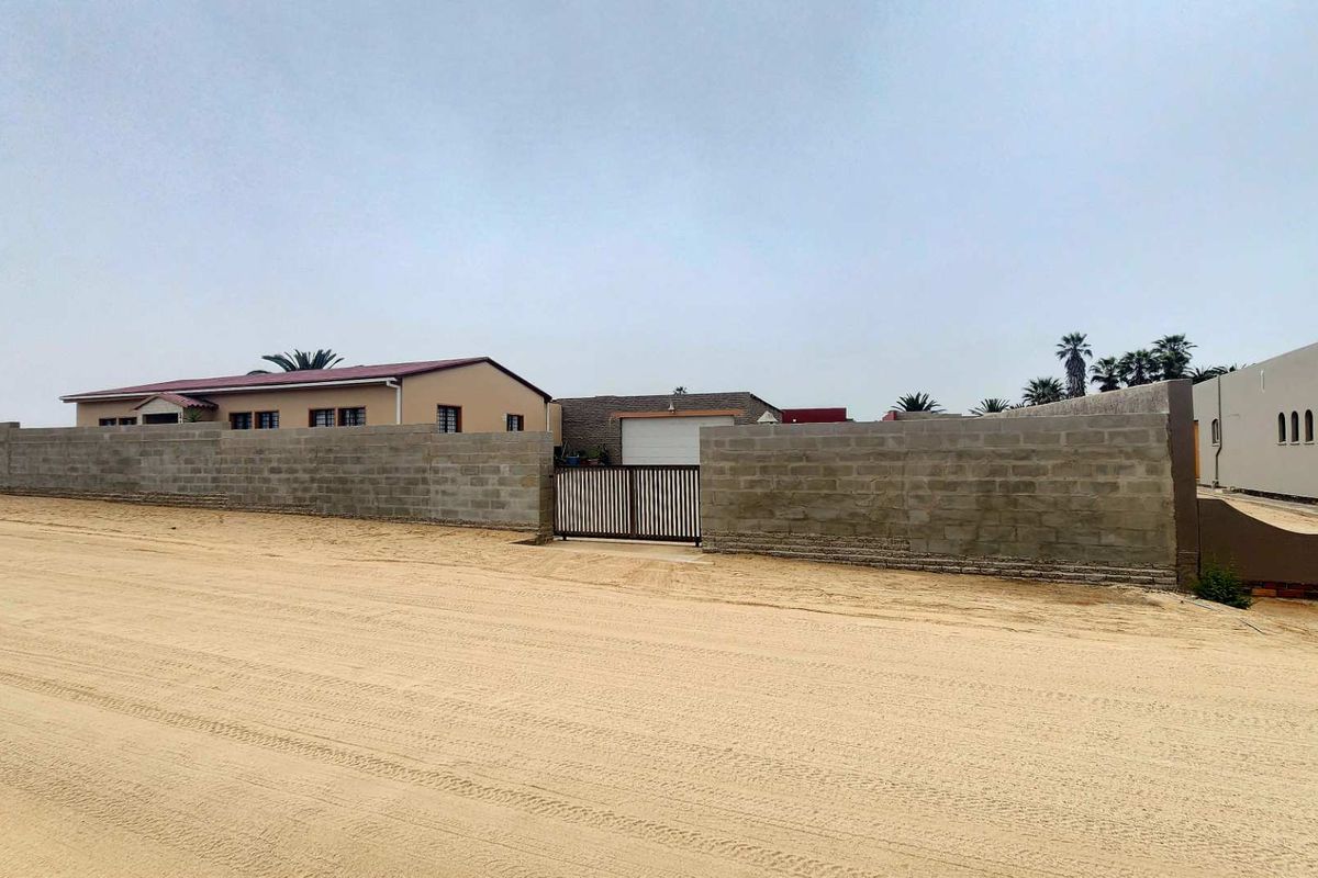 Front view of the property featuring a perimeter wall, metal gate, and garage access from a sandy road.