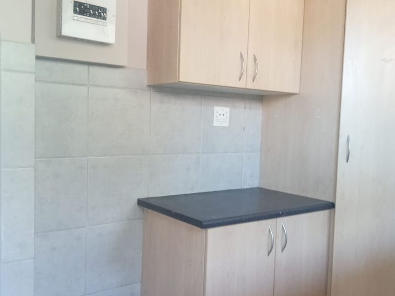 A compact kitchen area featuring light wood-finish cabinetry, a dark laminate countertop, and neutral tiled splashback with an integrated electrical distribution board.