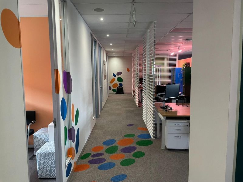 A modern office corridor featuring carpet flooring with decorative patterns, glass partitions, and slatted room dividers leading into an open-plan workspace.