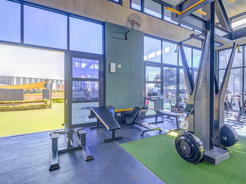 Modern gym facility featuring floor-to-ceiling windows, rubber flooring, artificial turf sections, and direct access to an outdoor area.