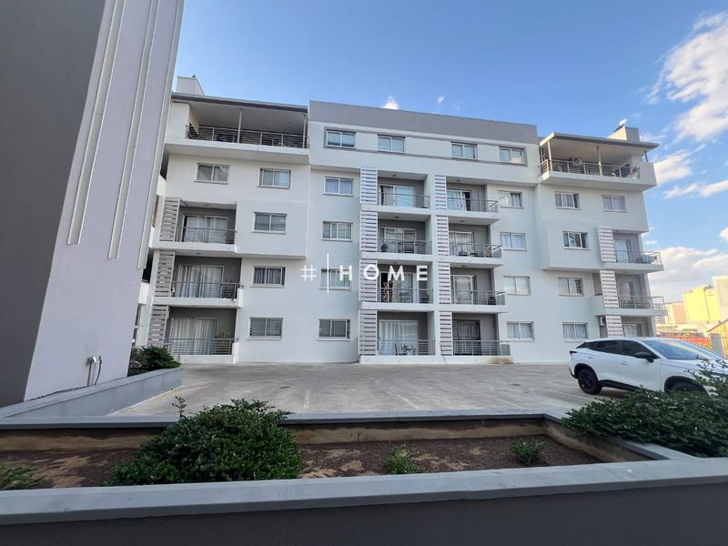 A modern multi-story apartment building featuring private balconies, sliding glass doors, and a paved parking area at the front entrance.