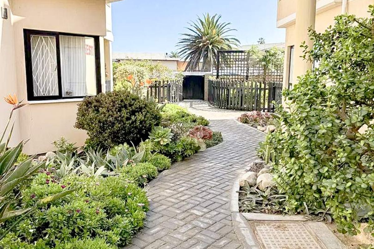 A paved walkway leads through a landscaped garden area between two apartment building sections, featuring established shrubs and succulents.