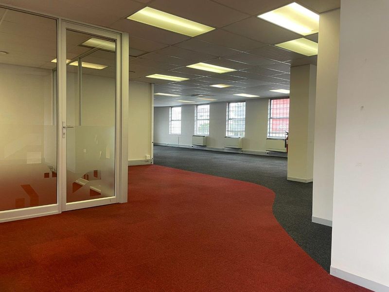 Spacious open-plan office area featuring glass partitioning, suspended ceilings with recessed lighting, and dual-tone carpet flooring with large windows.