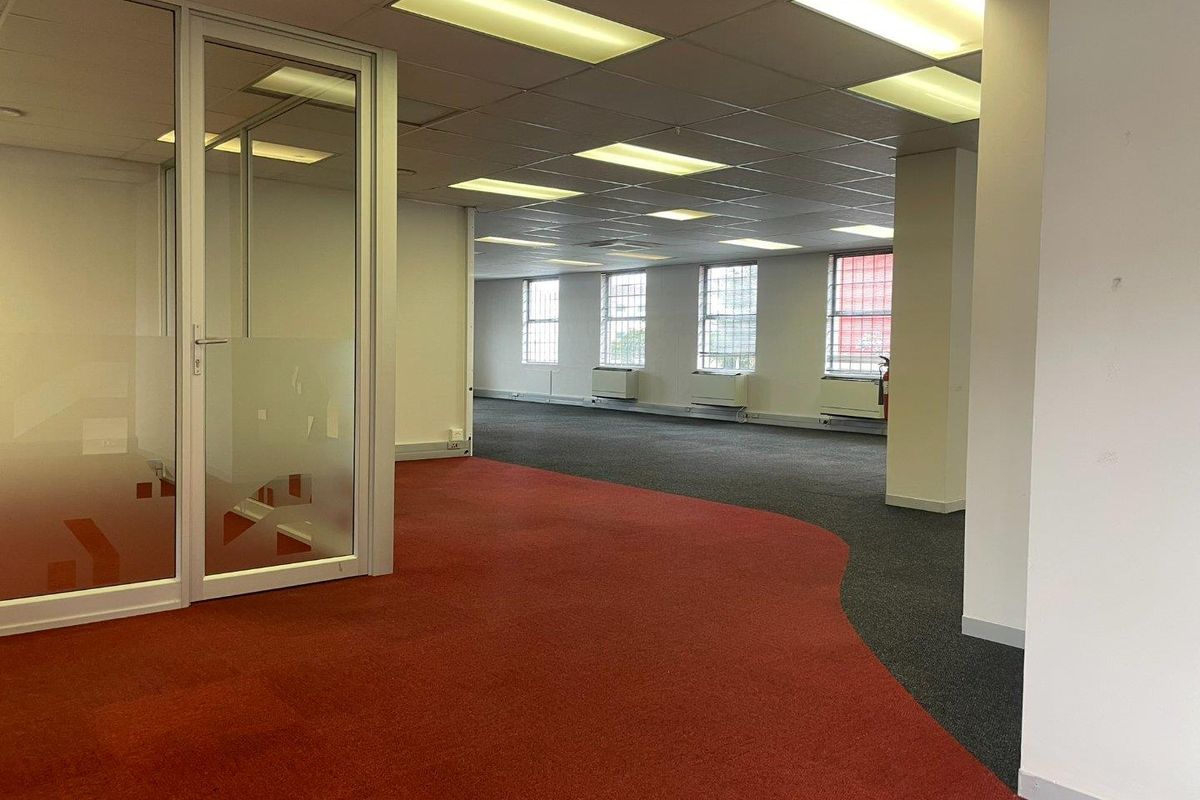 Spacious open-plan office area featuring glass partitioning, suspended ceilings with recessed lighting, and dual-tone carpet flooring with large windows.