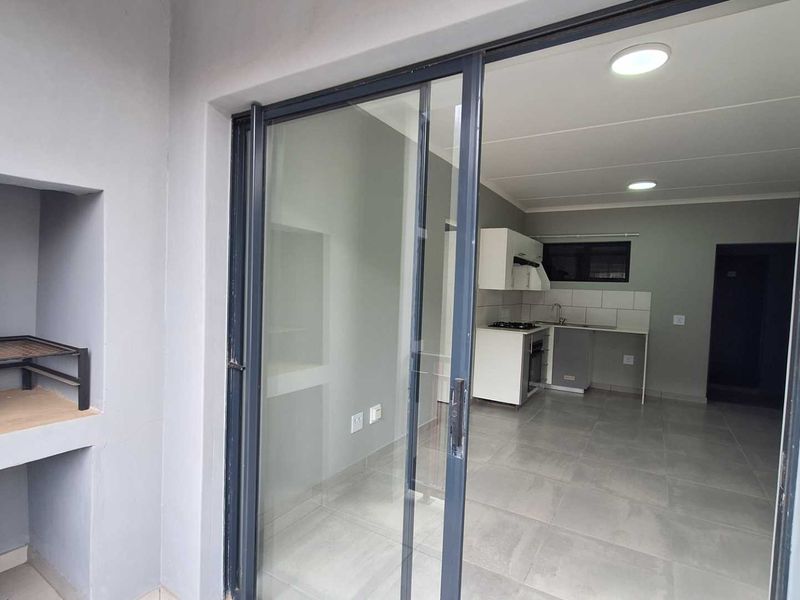 A covered patio featuring a built-in braai, leading through sliding glass doors into an open-plan kitchen area with modern tiled flooring.