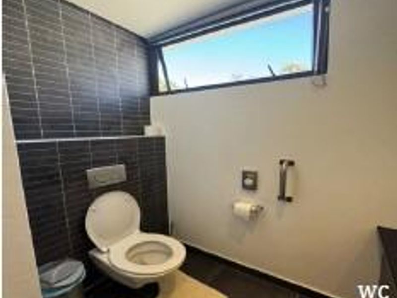 A modern, accessible office restroom featuring a wall-mounted toilet, dark tiled accent wall, and a high-level awning window for natural light.