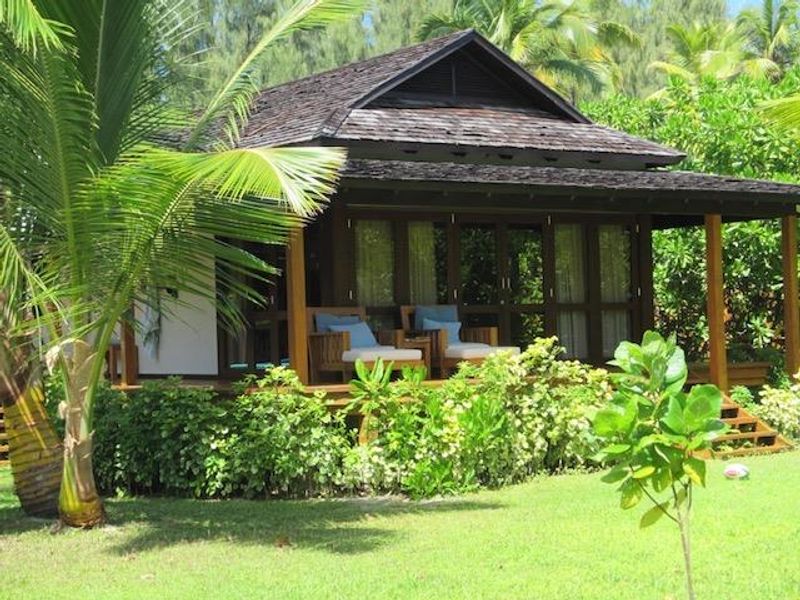 A charming villa exterior featuring a dark timber shingle roof, wrap-around wooden deck, and large glass sliding doors set within a lush tropical garden.