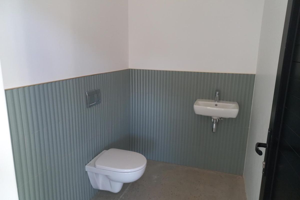 A modern, minimalist office restroom featuring a wall-mounted toilet, a compact wall-hung basin, and stylish sage green fluted wall tiling.