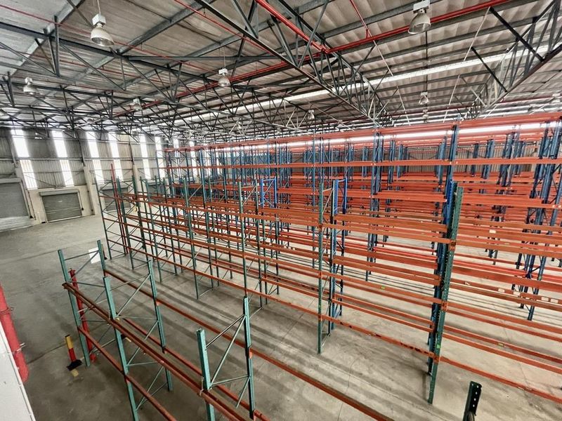 Spacious industrial warehouse featuring high-bay lighting, exposed steel roof trusses, concrete flooring, and extensive built-in industrial racking systems.