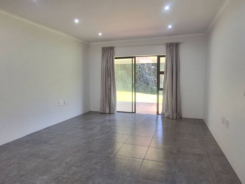 A bright, empty living room featuring modern grey floor tiles, recessed downlighting, and a sliding glass door providing access to the outdoor area.