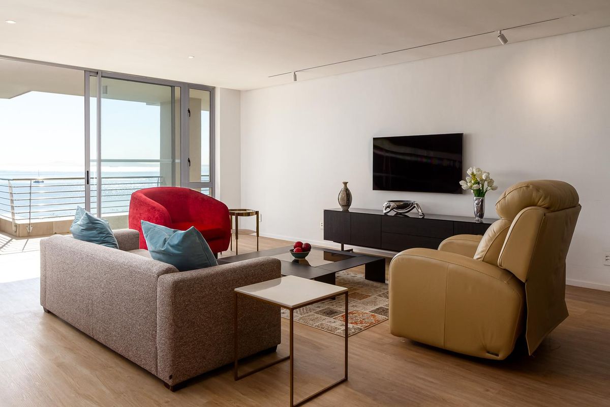 A bright and spacious living room featuring laminate flooring, track lighting, and large sliding glass doors that open onto a balcony with ocean views.