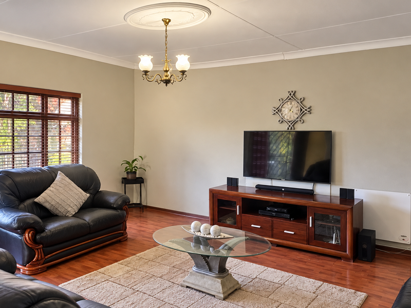 Spacious living room featuring warm laminate flooring, classic ceiling rose with chandelier, and large windows with timber frames.