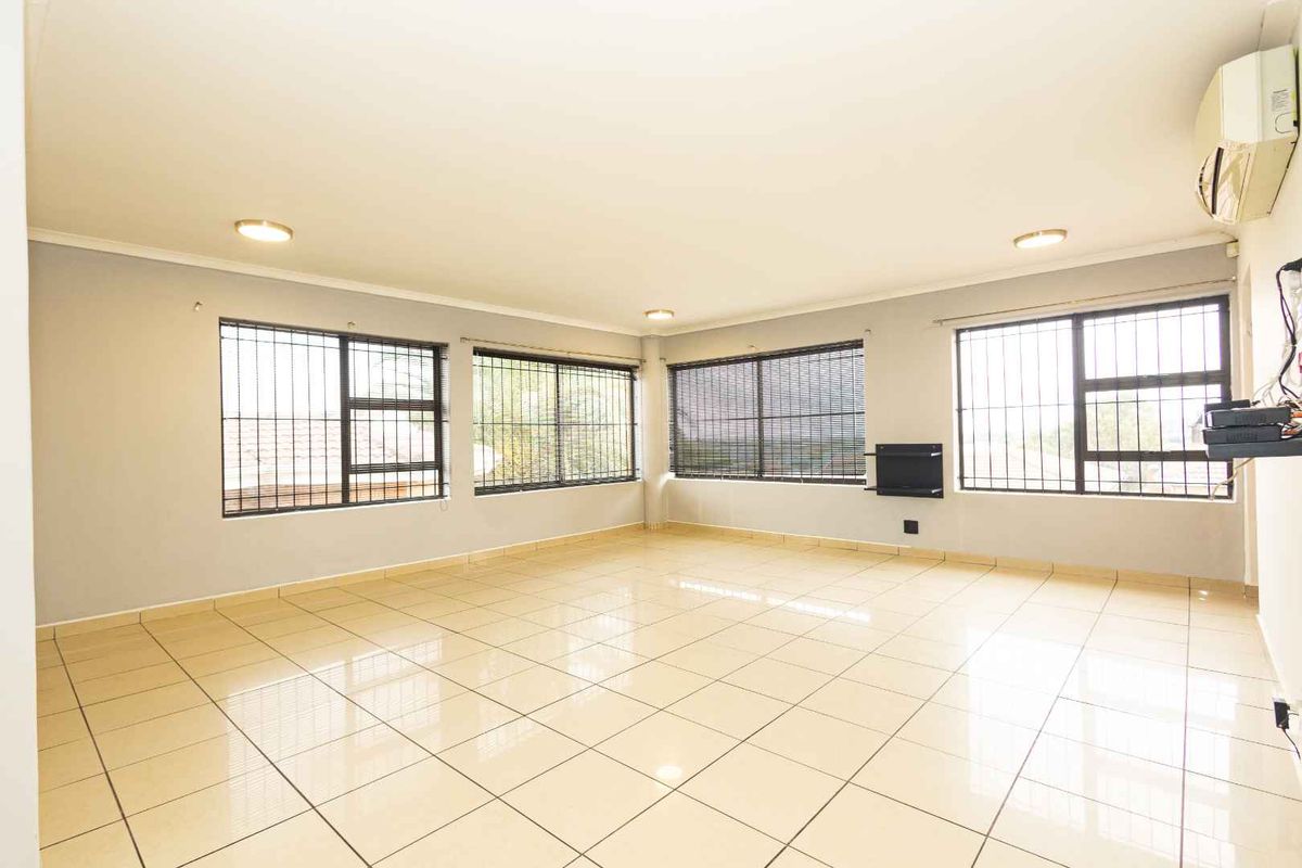 Spacious, light-filled living area featuring neutral tiled flooring, large windows with security bars, and a wall-mounted air conditioning unit.