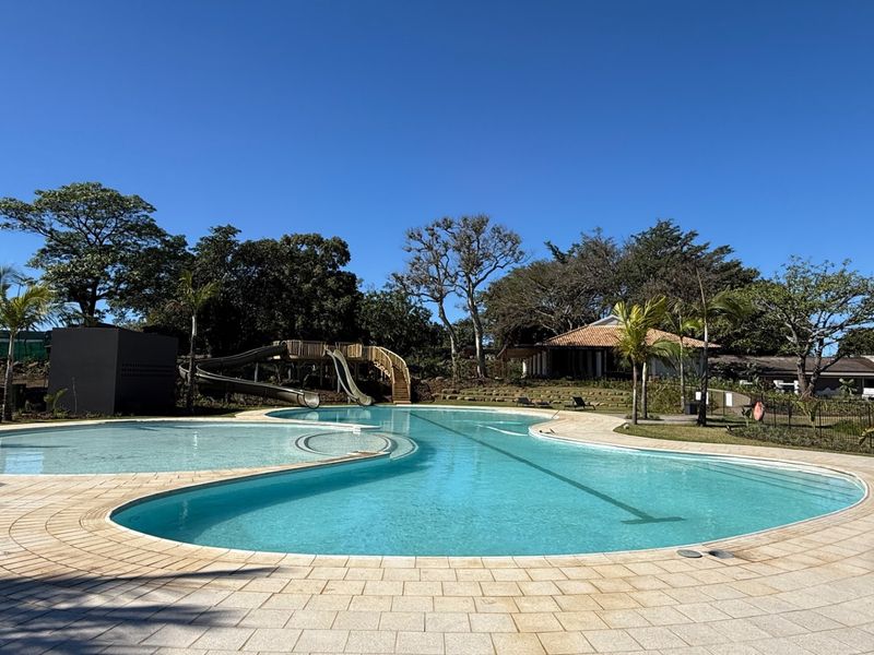 A well-maintained swimming pool area featuring a water slide, paved decking, and surrounding landscaped gardens in a peaceful outdoor setting.