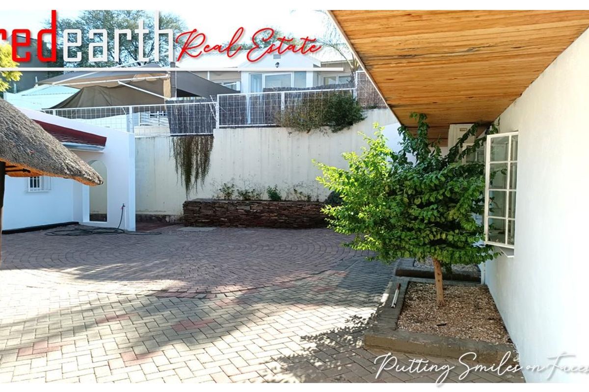 A paved rear courtyard area featuring a timber-clad overhang, a stone-built planter bed, and a thatched-roof structure.