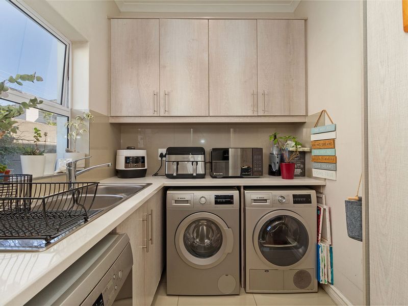 A functional laundry room featuring light wood-tone cabinetry, laminate countertops, a stainless steel sink, and dedicated spaces for under-counter appliances.