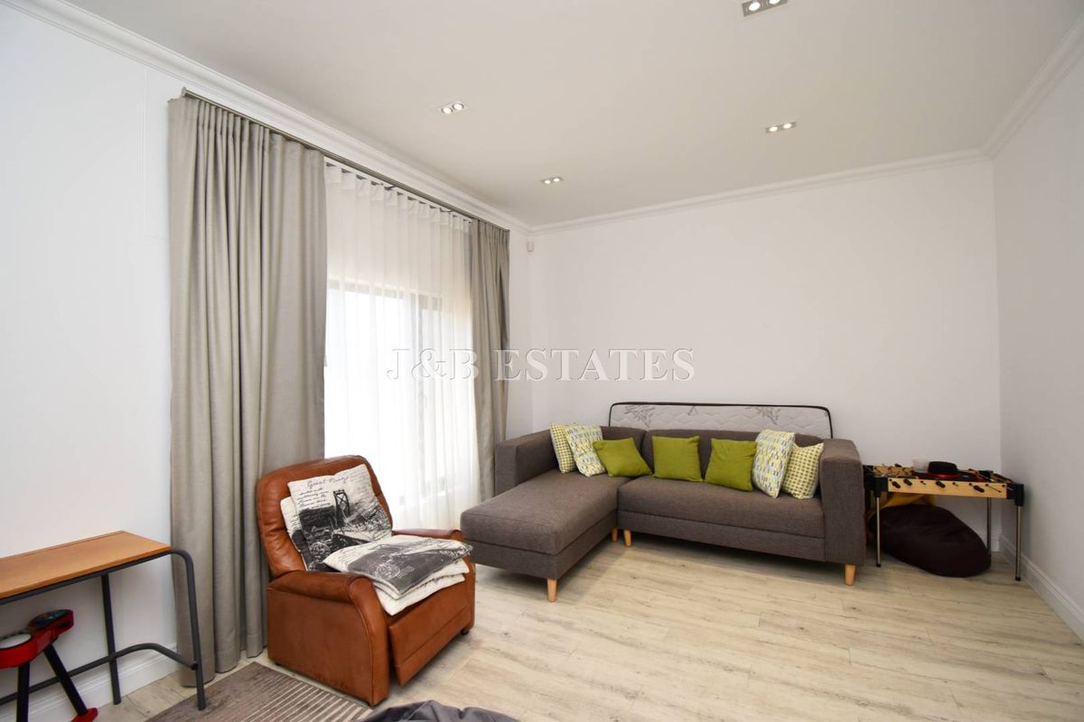 A bright and modern living room featuring light wood-look flooring, recessed ceiling lighting, and floor-to-ceiling curtains.