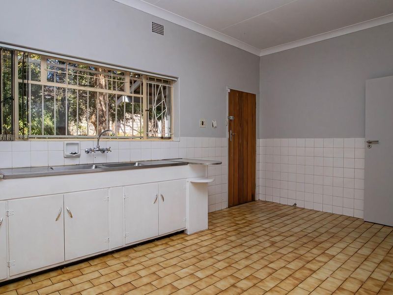 A spacious, empty kitchen featuring tiled flooring, built-in cabinetry, a stainless steel double sink, and a large window with security bars.
