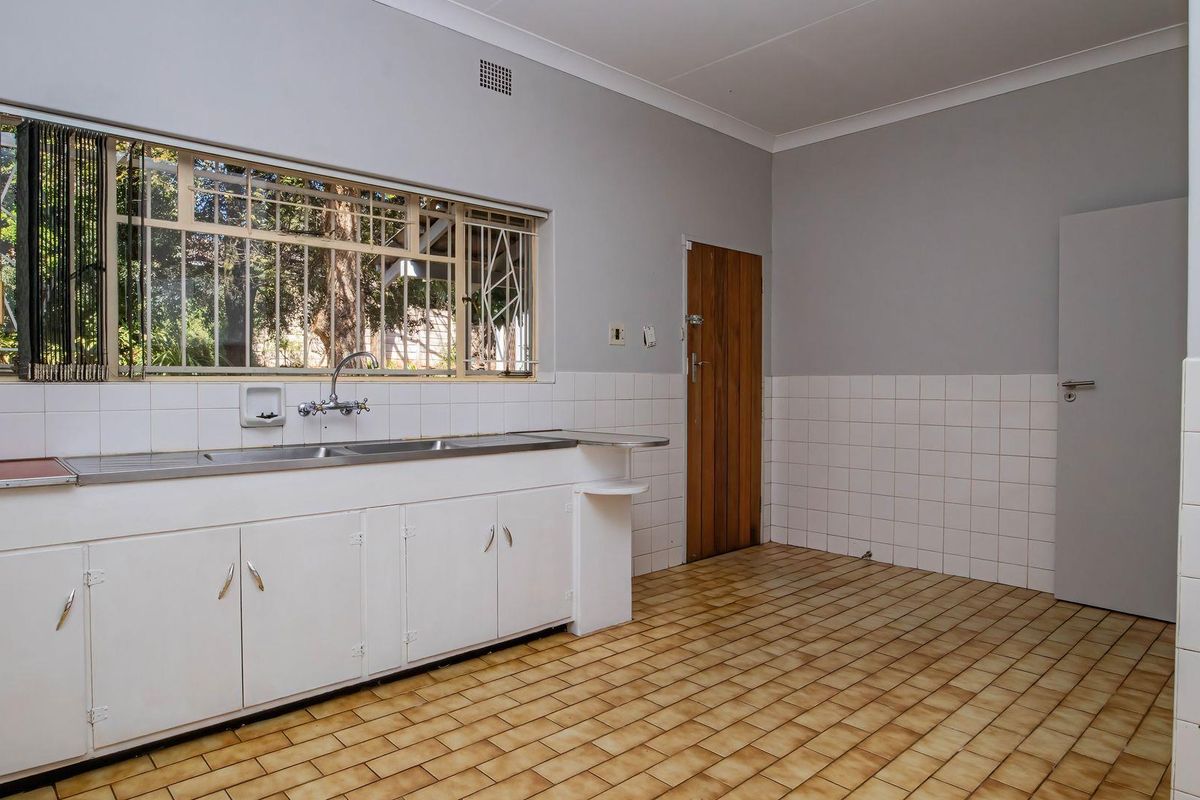 A spacious, empty kitchen featuring tiled flooring, built-in cabinetry, a stainless steel double sink, and a large window with security bars.